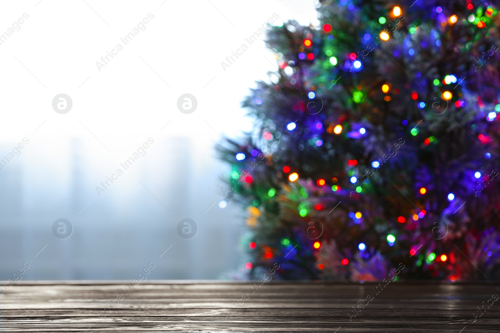 Photo of Blurred view of beautiful Christmas tree with colorful lights near window indoors, focus on wooden table. Space for text