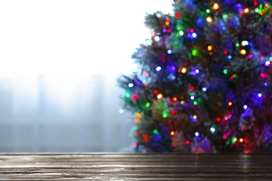 Photo of Blurred view of beautiful Christmas tree with colorful lights near window indoors, focus on wooden table. Space for text