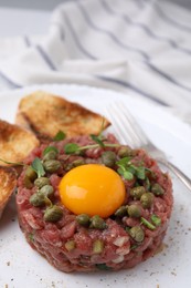 Photo of Tasty beef steak tartare served with yolk, capers, toasted bread and greens on plate, closeup