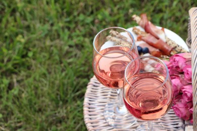 Photo of Glasses of delicious rose wine, food and flowers on picnic basket outdoors. Space for text
