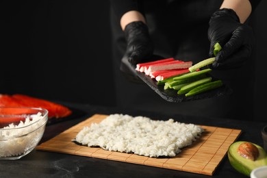 Photo of Chef in gloves putting cucumber onto unwrapped sushi roll at dark table, closeup