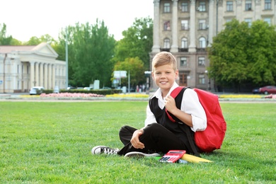 Cute boy with school stationery sitting on green lawn outdoors