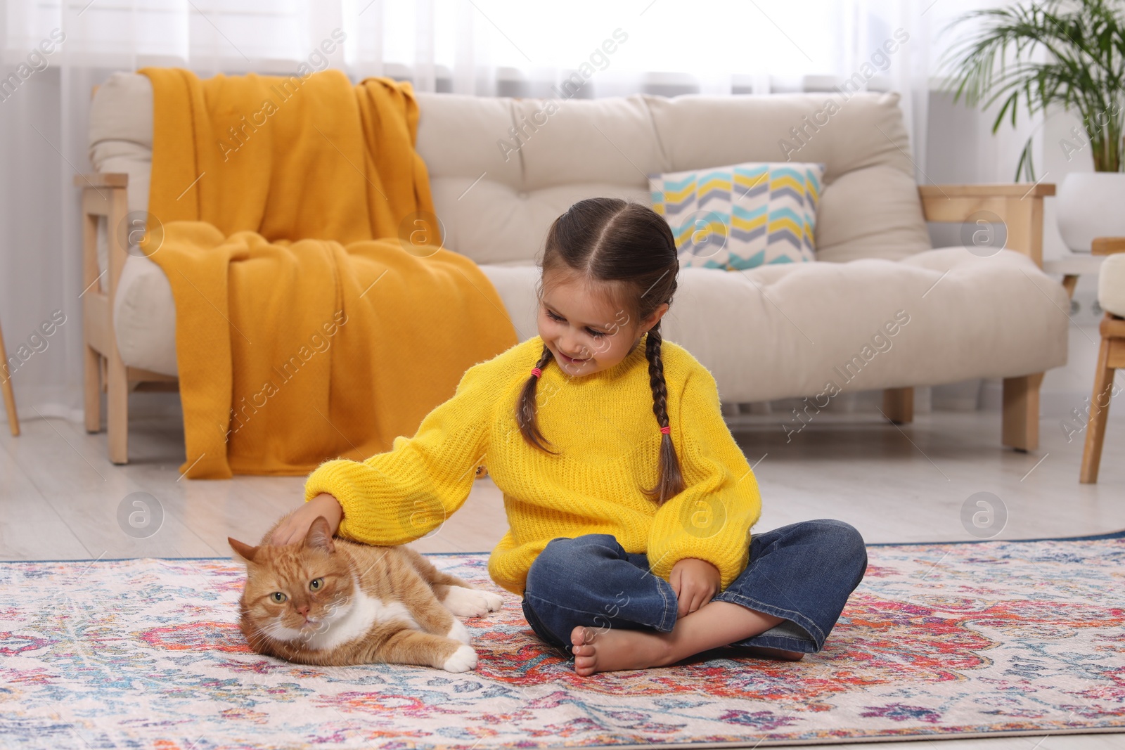 Photo of Smiling little girl petting cute ginger cat on carpet at home