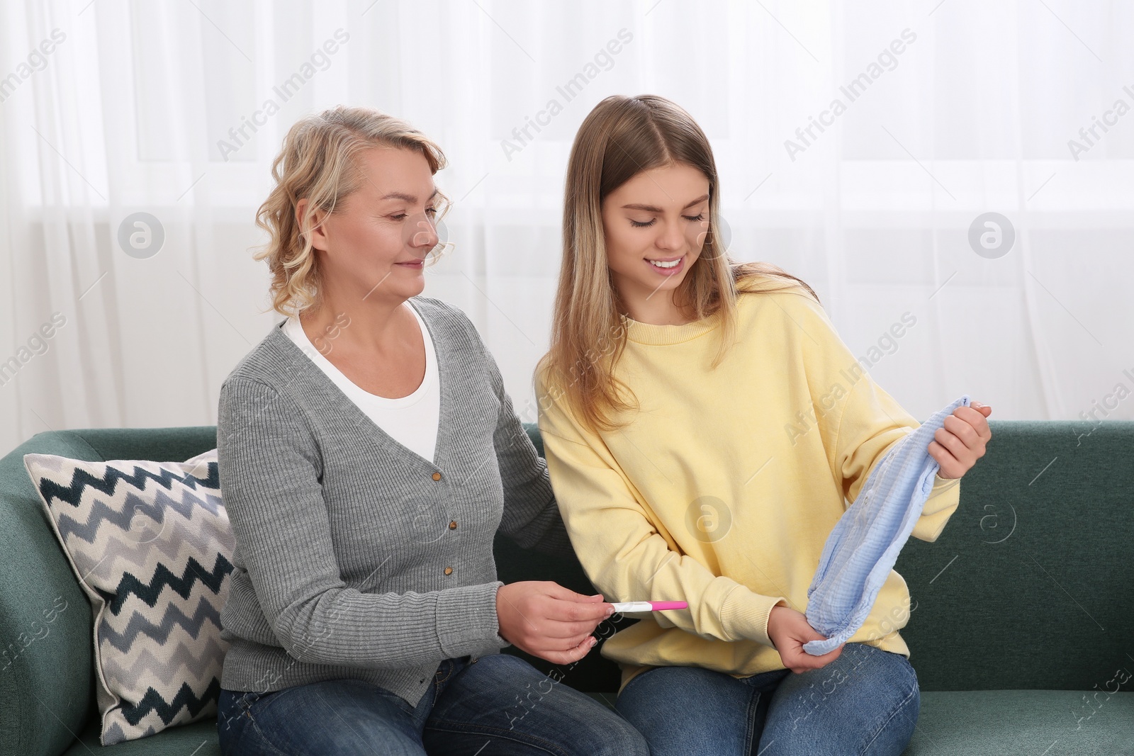 Photo of Young woman showing baby's bodysuit to her mother with pregnancy test at home. Grandparent reaction to future grandson