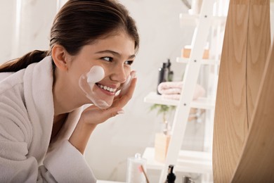 Photo of Beautiful teenage girl applying cleansing foam onto face in bathroom. Skin care cosmetic