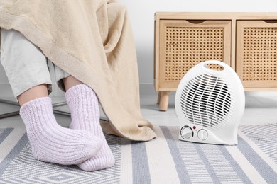 Photo of Woman warming feet near electric fan heater at home, closeup