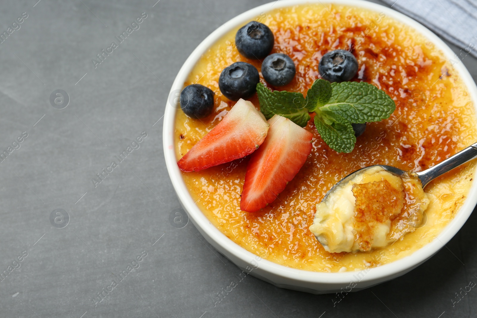 Photo of Delicious creme brulee with berries in bowl and spoon on grey table, closeup. Space for text