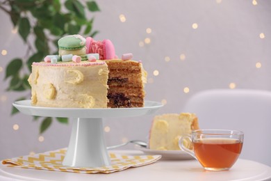 Delicious cake decorated with macarons and marshmallows and cup of tea on white table against blurred lights