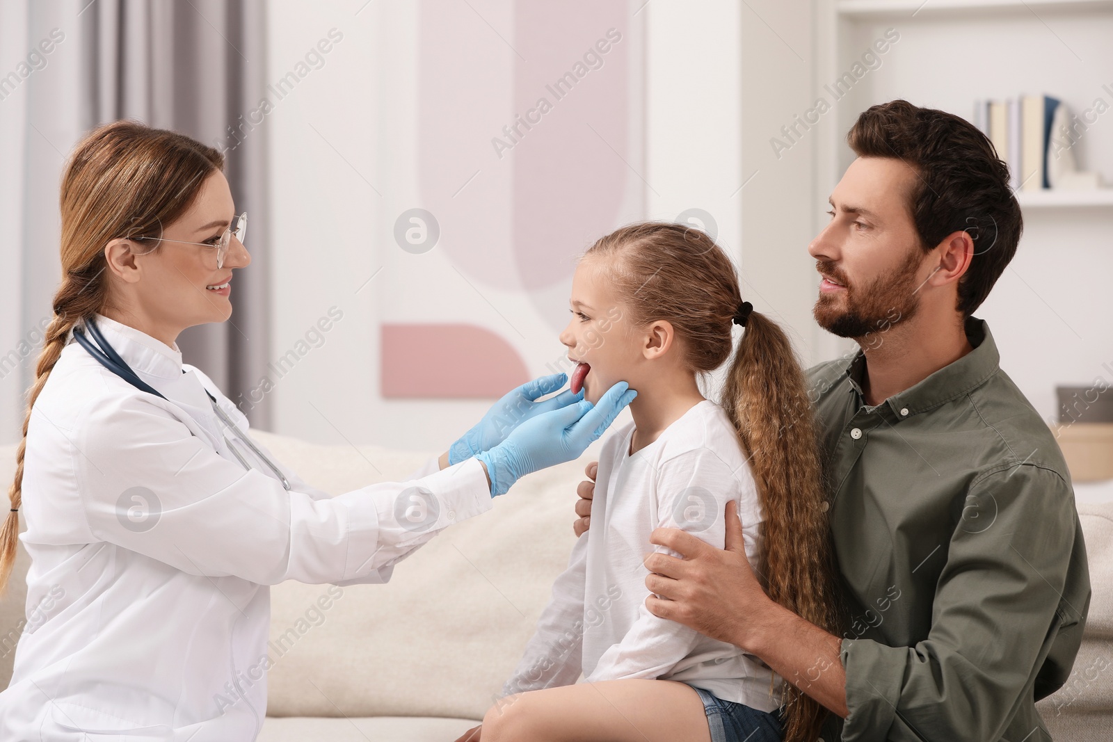 Photo of Smiling doctor examining girl`s oral cavity near her father indoors