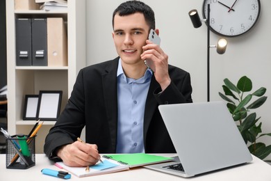 Photo of Man taking notes while talking on smartphone at table in office