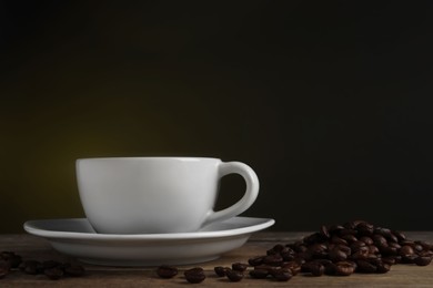 Photo of Cup of hot aromatic coffee and roasted beans on wooden table against dark background