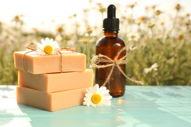 Photo of Bottle of chamomile essential oil and soap bars on light blue wooden table in field
