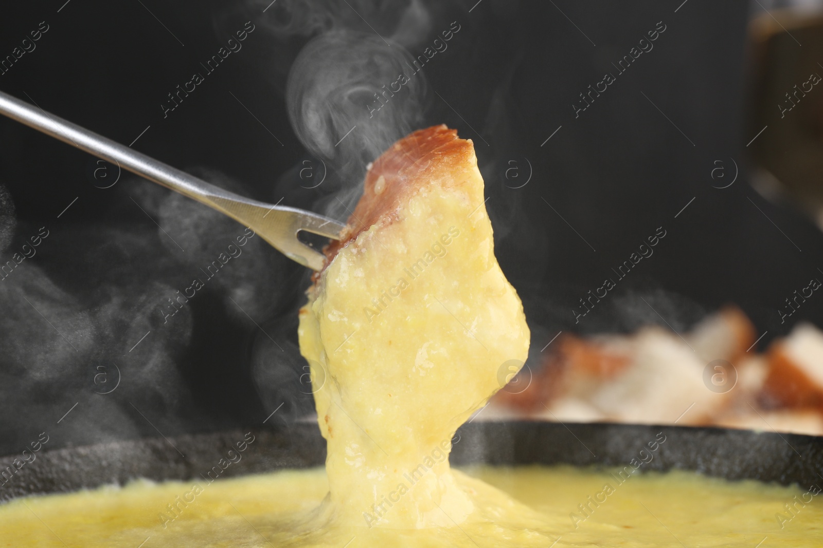 Image of Dipping piece of bread into fondue pot with tasty melted cheese against blurred background, closeup