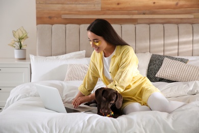 Young woman with eye patches working on laptop near her dog in bedroom. Home office concept