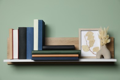 Wooden shelf with many hardcover books, picture and dry flowers on light green wall