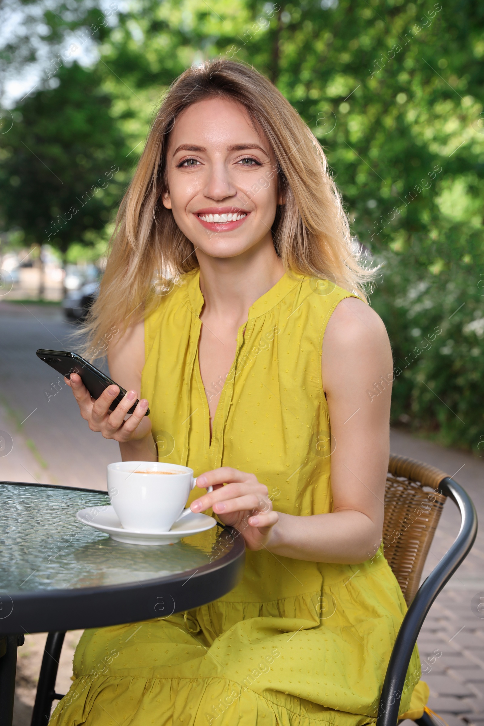 Photo of Happy young woman with cup of coffee and smartphone enjoying early morning in outdoor cafe