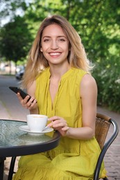 Photo of Happy young woman with cup of coffee and smartphone enjoying early morning in outdoor cafe