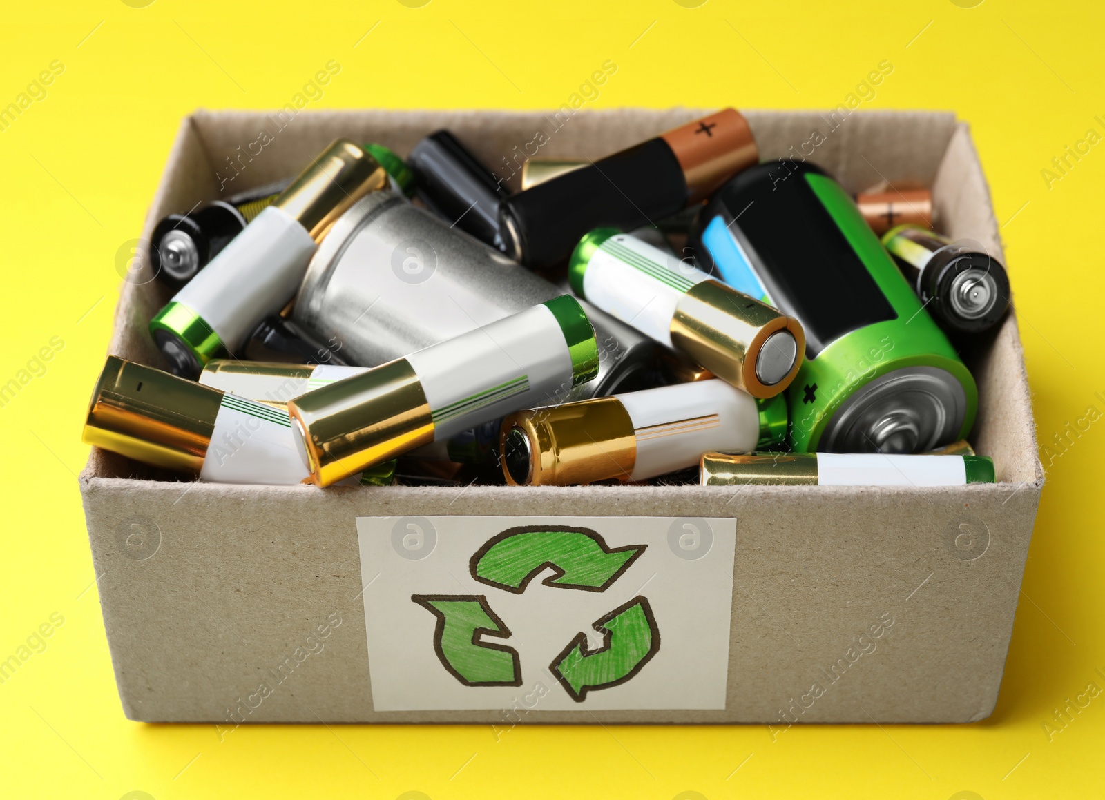 Image of Used batteries in cardboard box with recycling symbol on yellow background, closeup