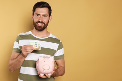 Photo of Happy man putting money into piggy bank on beige background, space for text