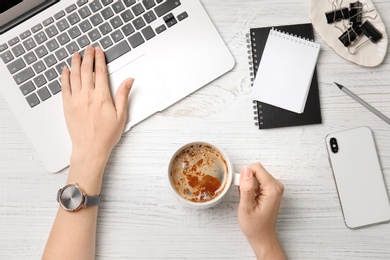 Young woman with cup of delicious coffee using laptop at table, top view