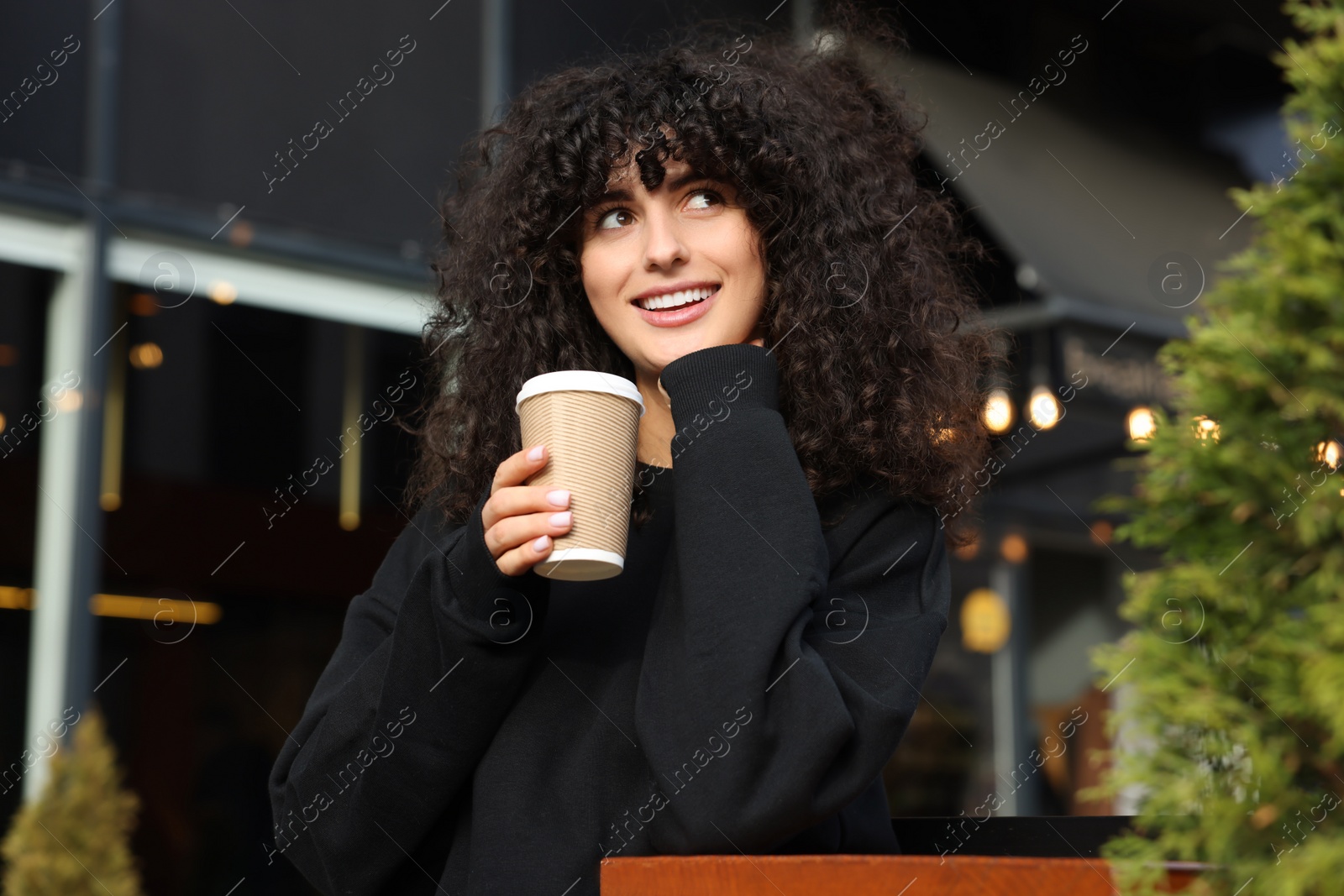 Photo of Happy young woman in stylish black sweater with cup of coffee at wooden table outdoors