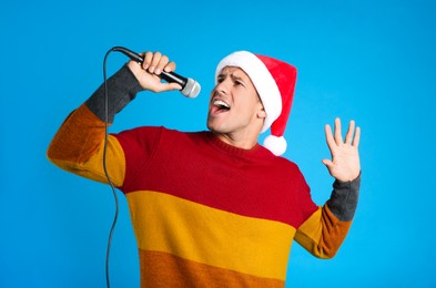 Emotional man in Santa Claus hat singing with microphone on blue background. Christmas music