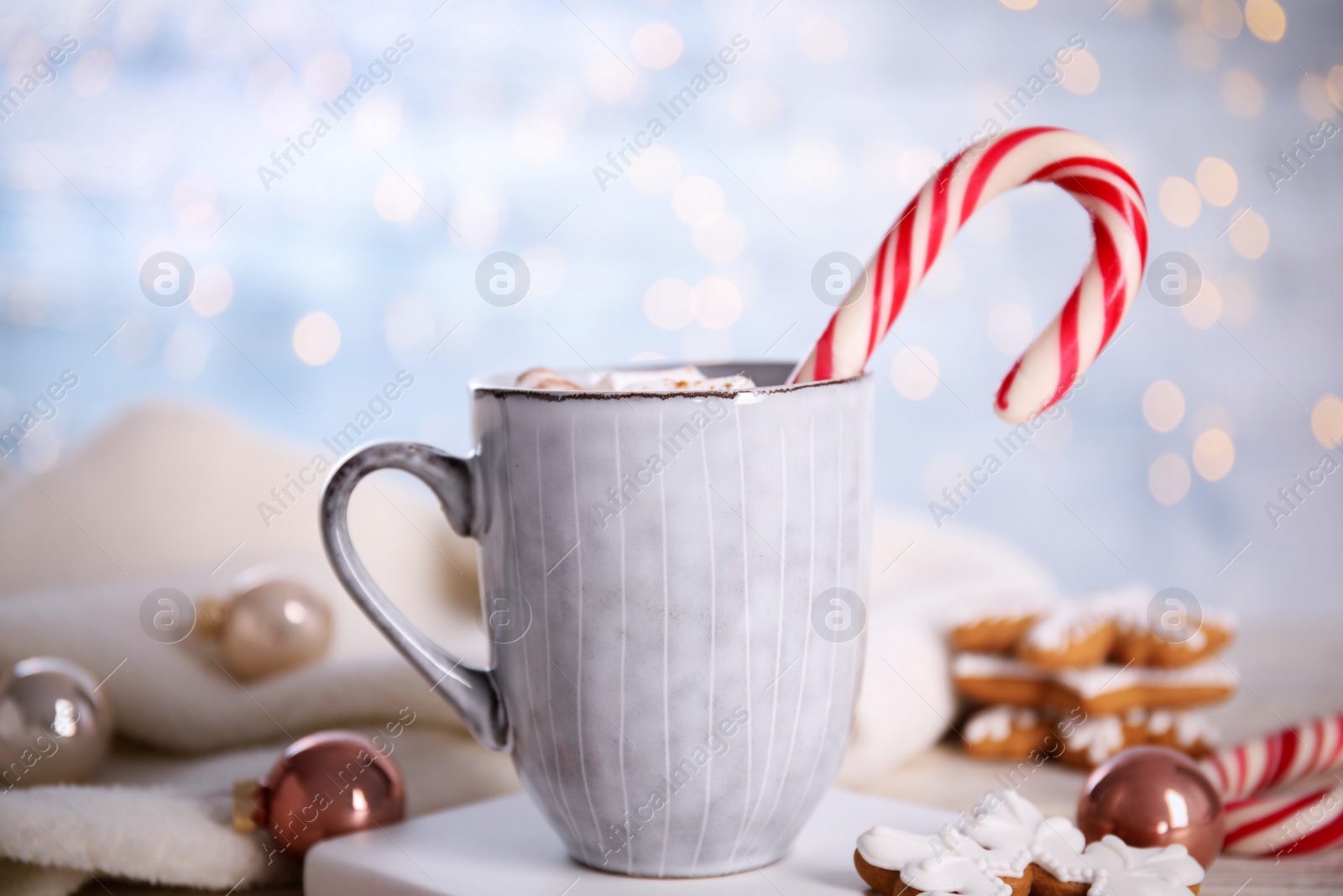 Photo of Cup of tasty cocoa with marshmallows and Christmas candy cane on white stand against blurred festive lights