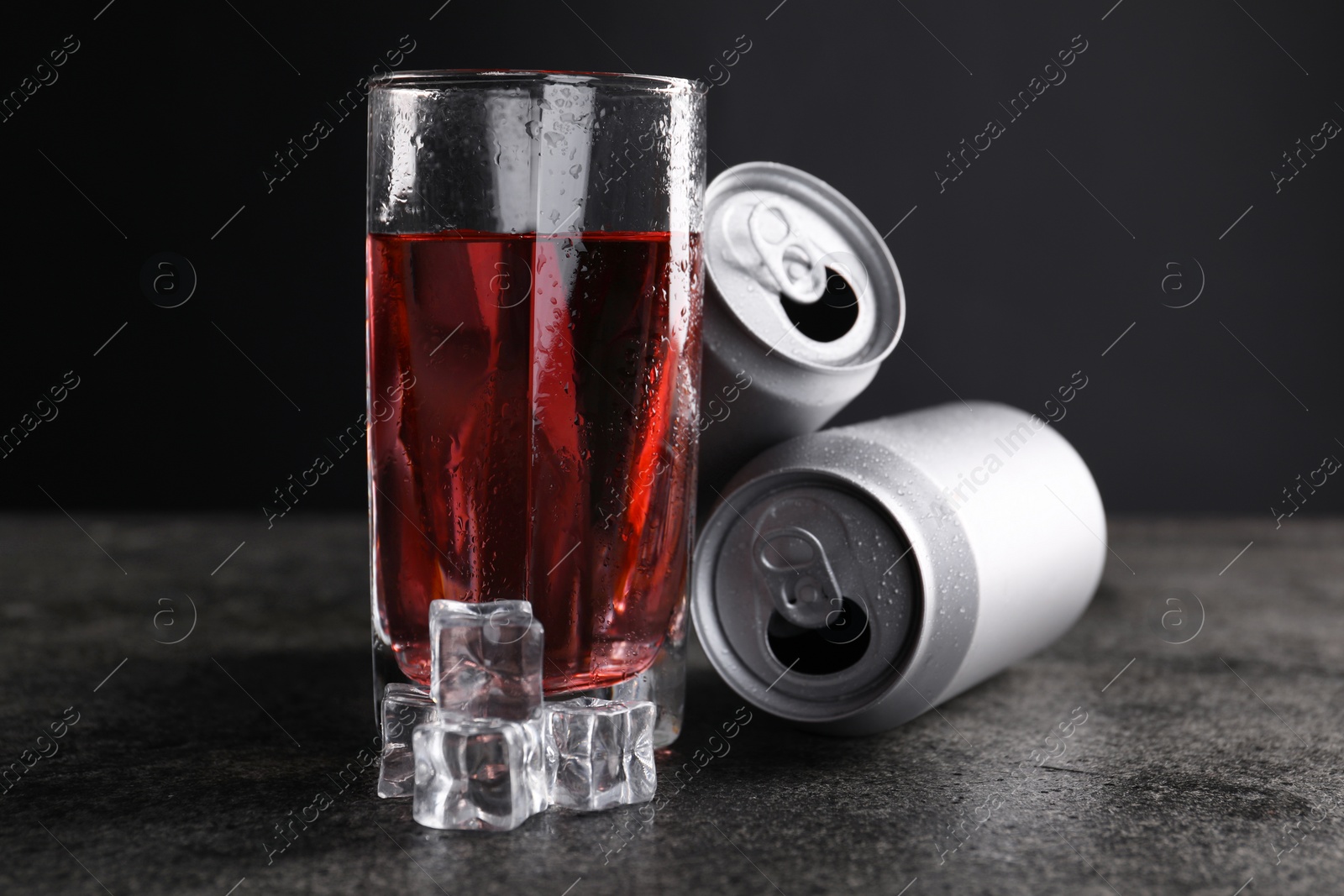 Photo of Energy drink in glass, aluminium cans and ice cubes on grey table, closeup