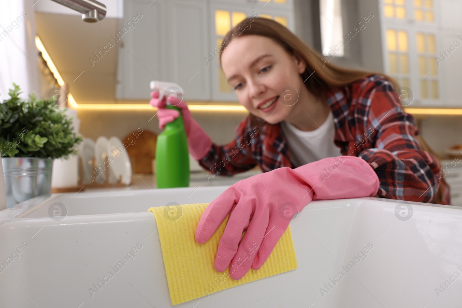 Photo of Woman with spray bottle and microfiber cloth cleaning sink in kitchen, selective focus