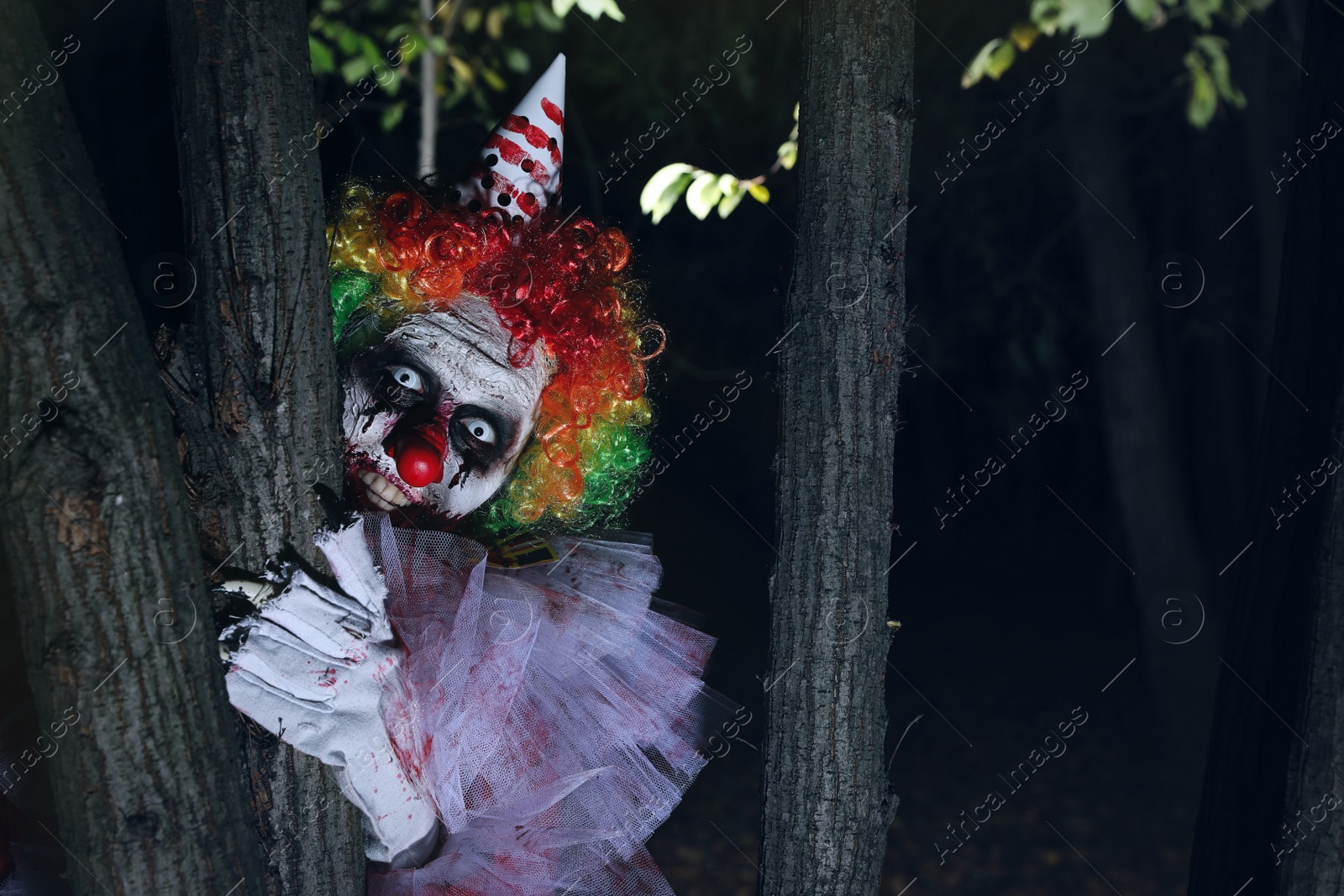 Photo of Terrifying clown hiding behind trees outdoors at night. Halloween party costume