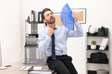 Bearded businessman waving blue hand fan to cool himself in office