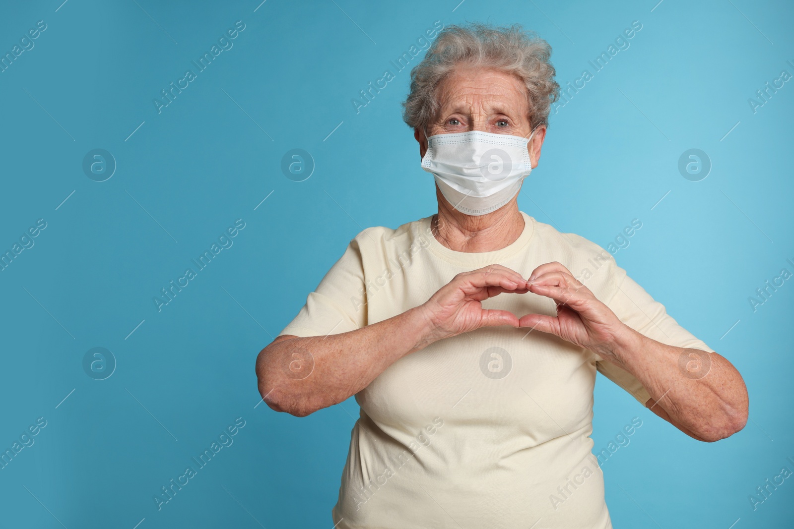 Photo of Elderly woman in medical mask making heart with her hands on light blue background, space for text