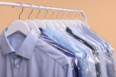 Photo of Dry-cleaning service. Many different clothes in plastic bags hanging on rack against beige background, closeup