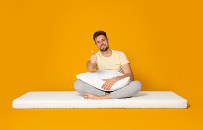 Photo of Smiling man sitting on soft mattress and showing thumb up against orange background