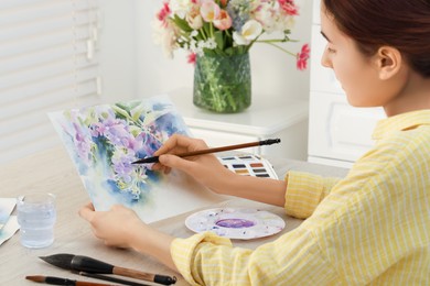 Photo of Woman painting flowers with watercolor at white wooden table indoors. Creative artwork