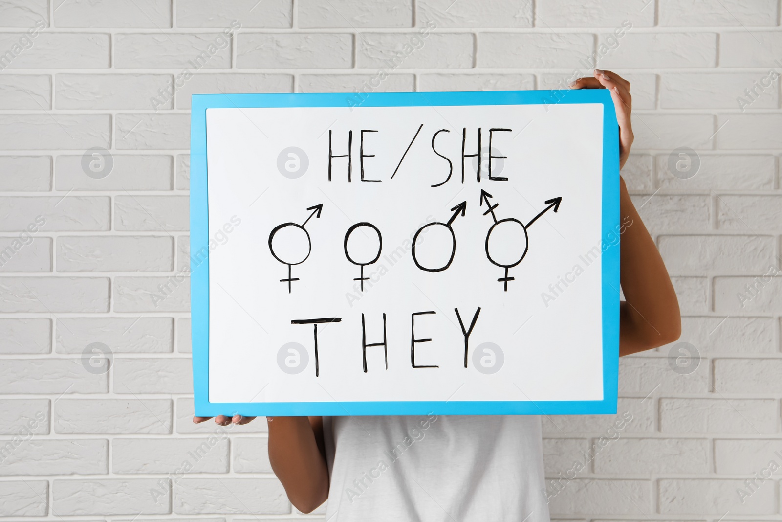 Photo of Woman holding sign with gender pronouns and symbols near white brick wall