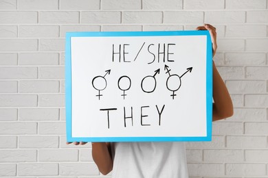 Photo of Woman holding sign with gender pronouns and symbols near white brick wall