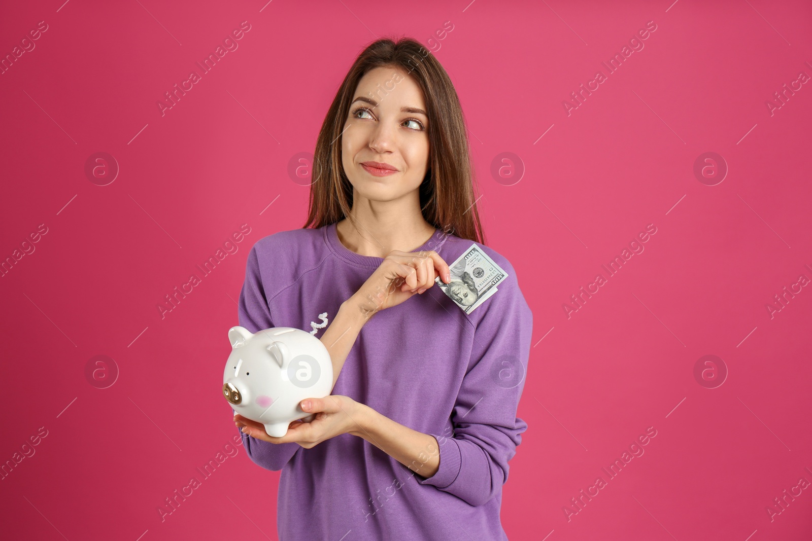 Photo of Happy young woman putting money into piggy bank on pink background