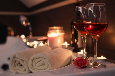 Photo of Glasses of wine, towels and rose on tub in bathroom, space for text. Romantic atmosphere