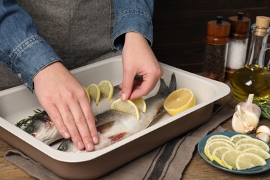 Photo of Woman putting pieces of lemon in raw sea bass fish at wooden table, closeup