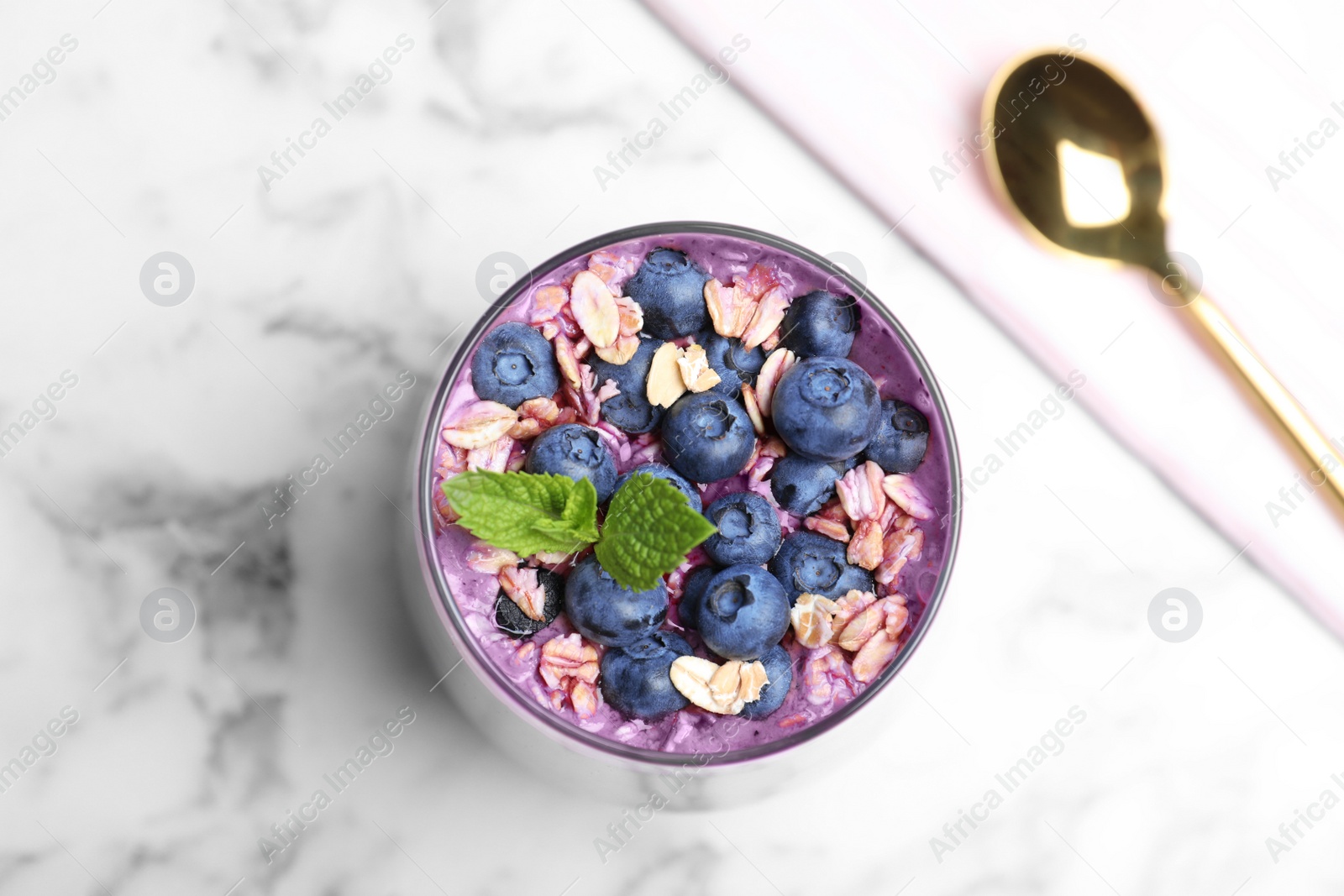 Photo of Glass of delicious blueberry smoothie with granola, mint and spoon on white marble table, flat lay