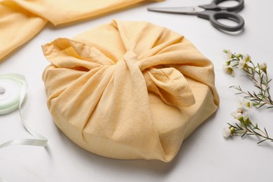 Photo of Furoshiki. Things packed in fabric, scissors, ribbon and flowers on white table, closeup