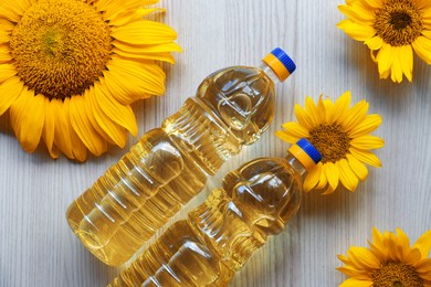 Photo of Bottles of sunflower oil and flowers on light wooden table, flat lay
