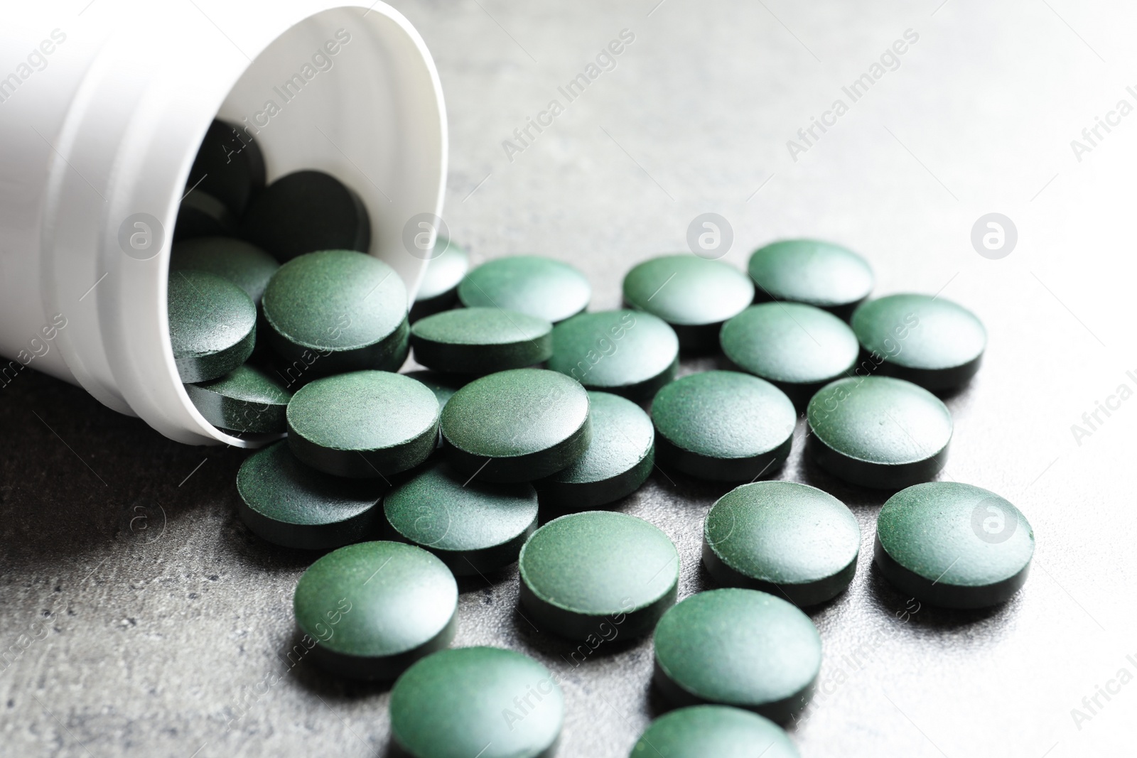 Photo of Plastic container with green spirulina pills on grey table, closeup