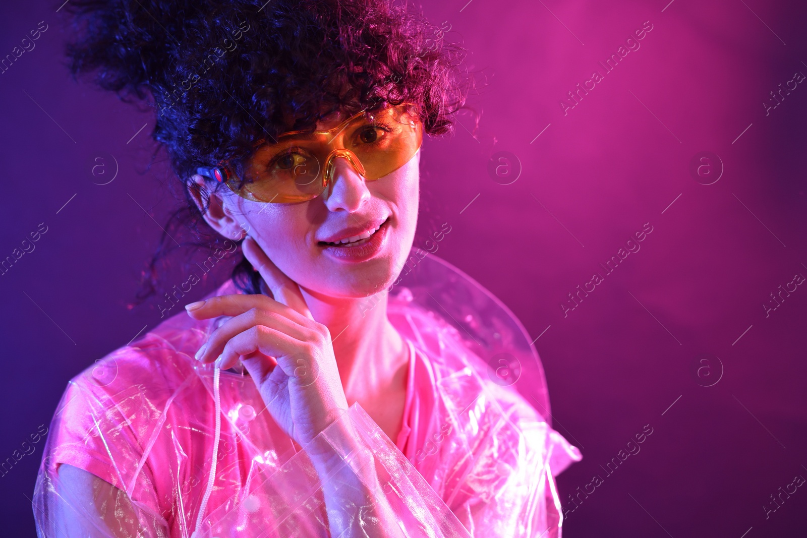 Photo of Beautiful young woman in transparent coat and sunglasses posing on color background in neon lights. Space for text