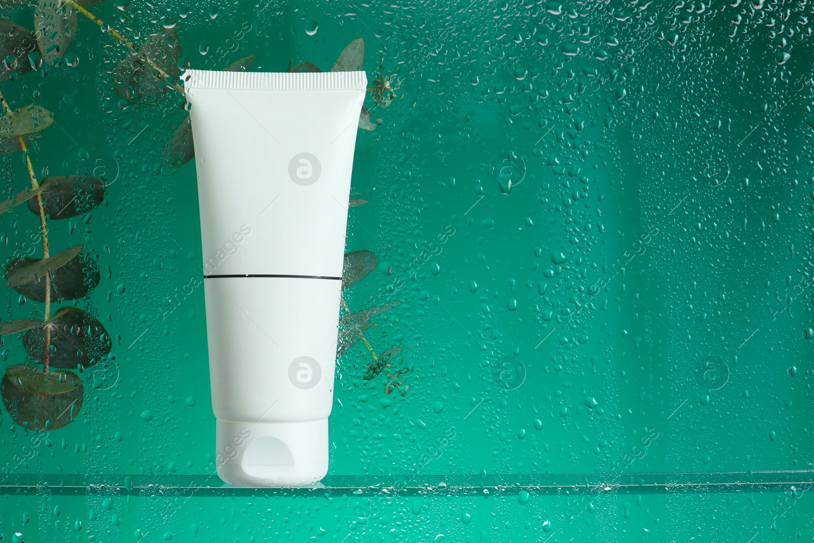 Photo of Tube with moisturizing cream and eucalyptus branches on green background, view through wet glass. Space for text