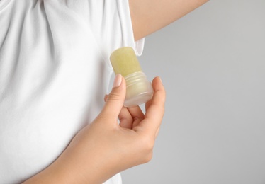 Photo of Young woman holding natural crystal alum deodorant near armpit on light grey background, closeup