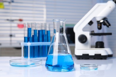 Photo of Laboratory analysis. Different glassware with blue liquid and microscope on white table indoors