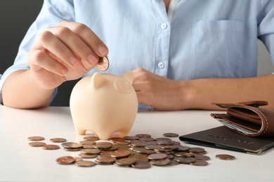 Photo of Young woman putting coin into piggy bank at wooden table, closeup view