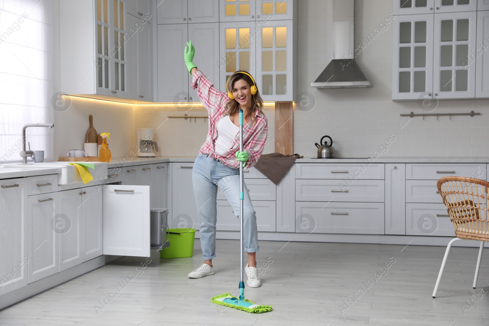 Photo of Beautiful young woman with headphones singing while cleaning kitchen